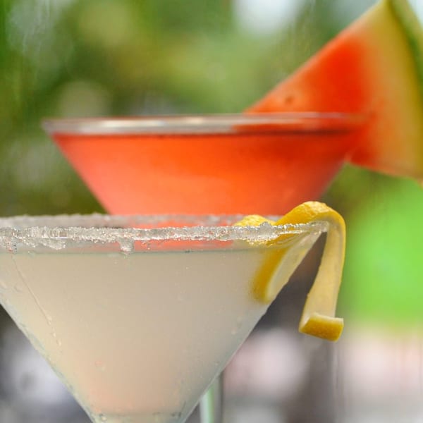 Two cocktails, one white with a lemon twist and sugar rim, one orange with a watermelon slice.