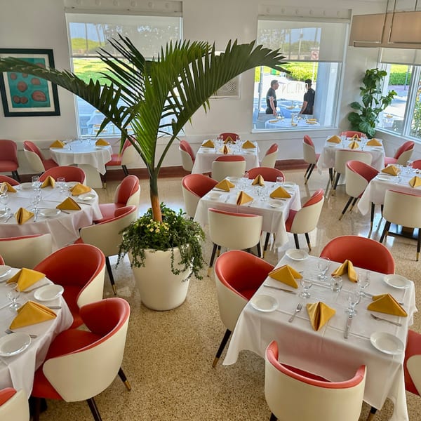 Bright Avalon By Day dining room with white-clothed tables, red chairs, and large windows overlooking greenery.