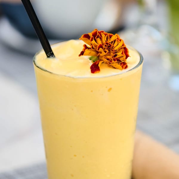 A vibrant yellow mango smoothie in a tall glass, garnished with an orange and red flower.
