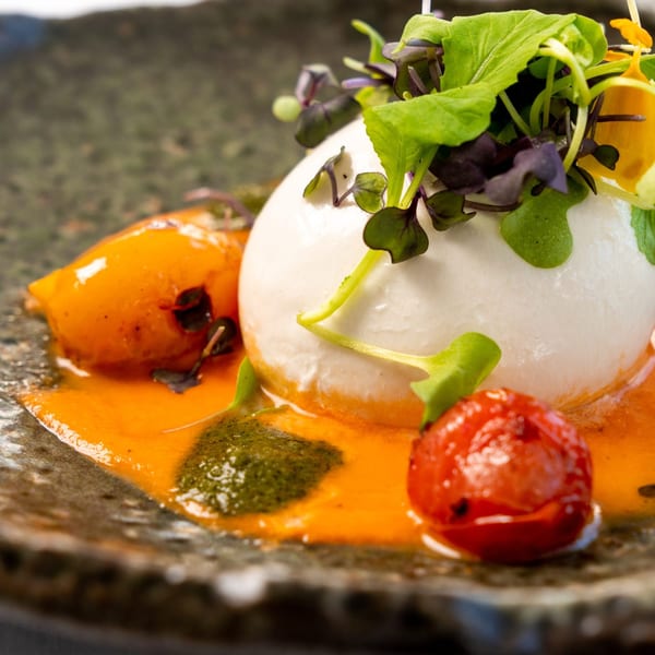 Burrata cheese with roasted tomatoes, orange sauce, pesto, and microgreens on a dark plate.