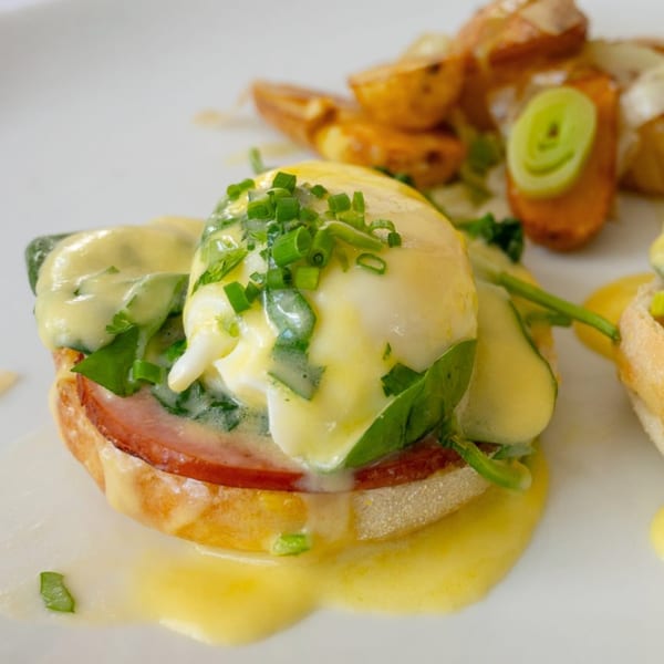 Eggs Benedict with hollandaise sauce, chives, spinach, ham, and roasted potatoes on a white plate.