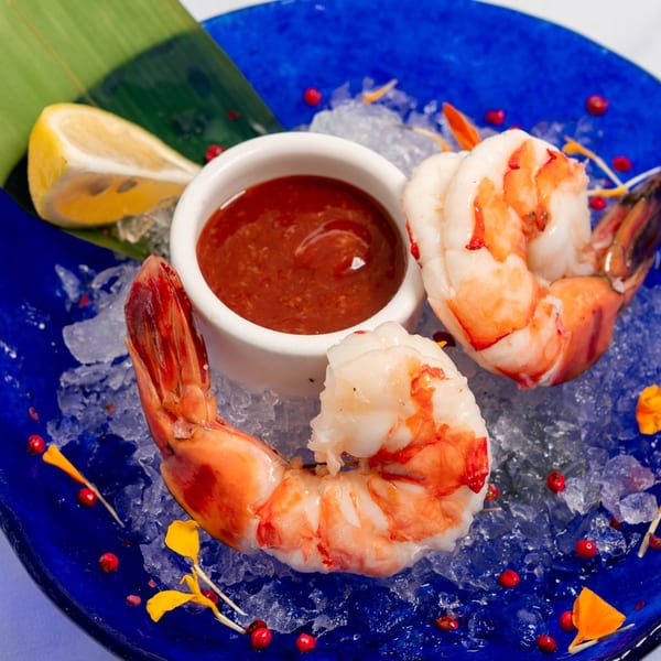 Shrimp cocktail with sauce and lemon on a bed of ice, garnished with red peppercorns.