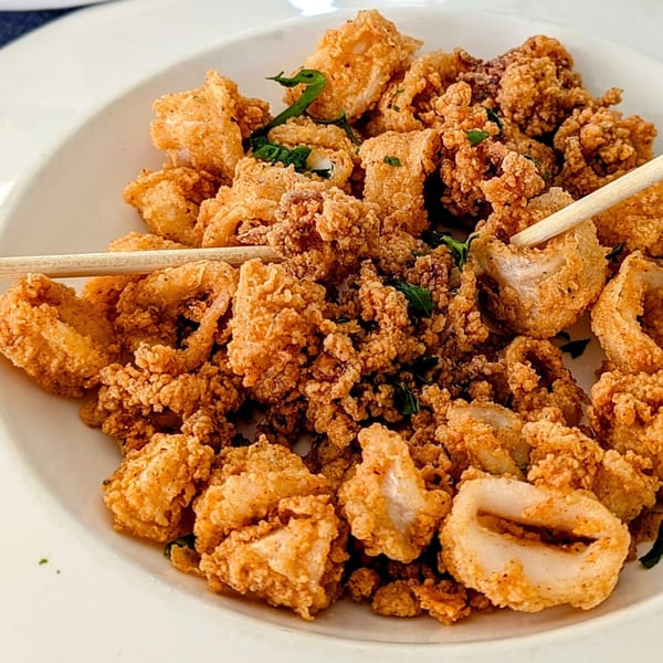 Close-up of a white bowl filled with golden-brown fried calamari, garnished with green herbs and two wooden skewers.