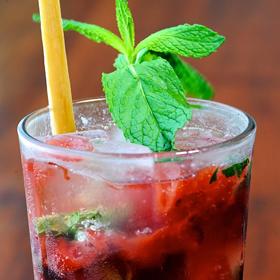 Close-up of a vibrant red cocktail with ice, fresh mint, and a yellow straw.
