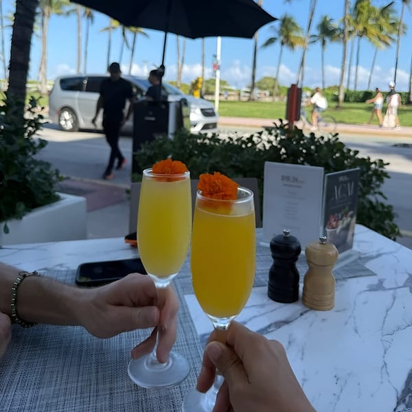 Two hands holding mimosa glasses with orange flower garnishes at an outdoor Avalon By Day table.