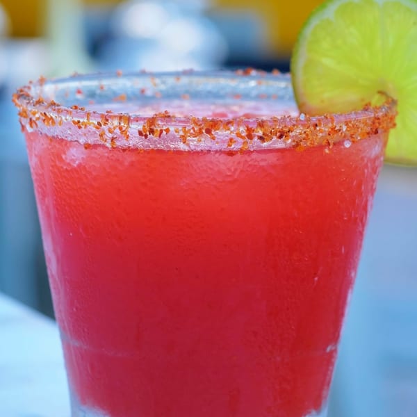 A vibrant red frozen drink with a chili-salt rim and a lime wedge garnish.