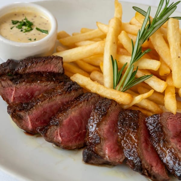Sliced steak with a side of golden french fries, a small dish of sauce, and a rosemary sprig.