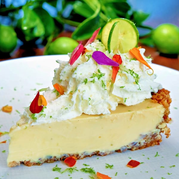 Slice of key lime pie with whipped cream, lime zest, a lime slice, and edible flower petals.