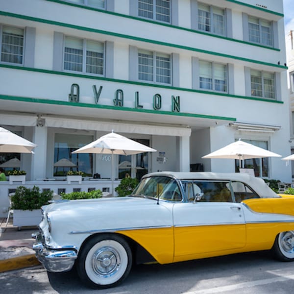 A vintage yellow and white car parked outside the Avalon Hotel, with outdoor dining umbrellas visible.